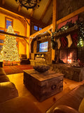 A cozy rustic lodge living room decorated for Christmas, featuring a glowing fireplace with stockings and a tree in the background, a rustic trunk coffee table in the foreground, and warm wood beams throughout.